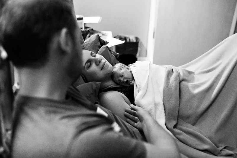 Image of a mother with her newborn resting on her chest while her partner strokes her arm.