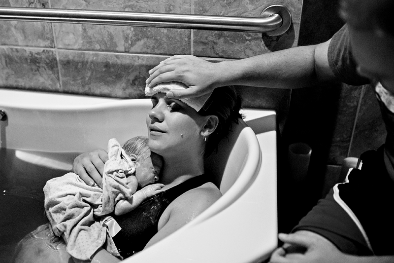 Image of a mother in a birthing pool with her newborn baby and her partner holding a cloth to her forehead.